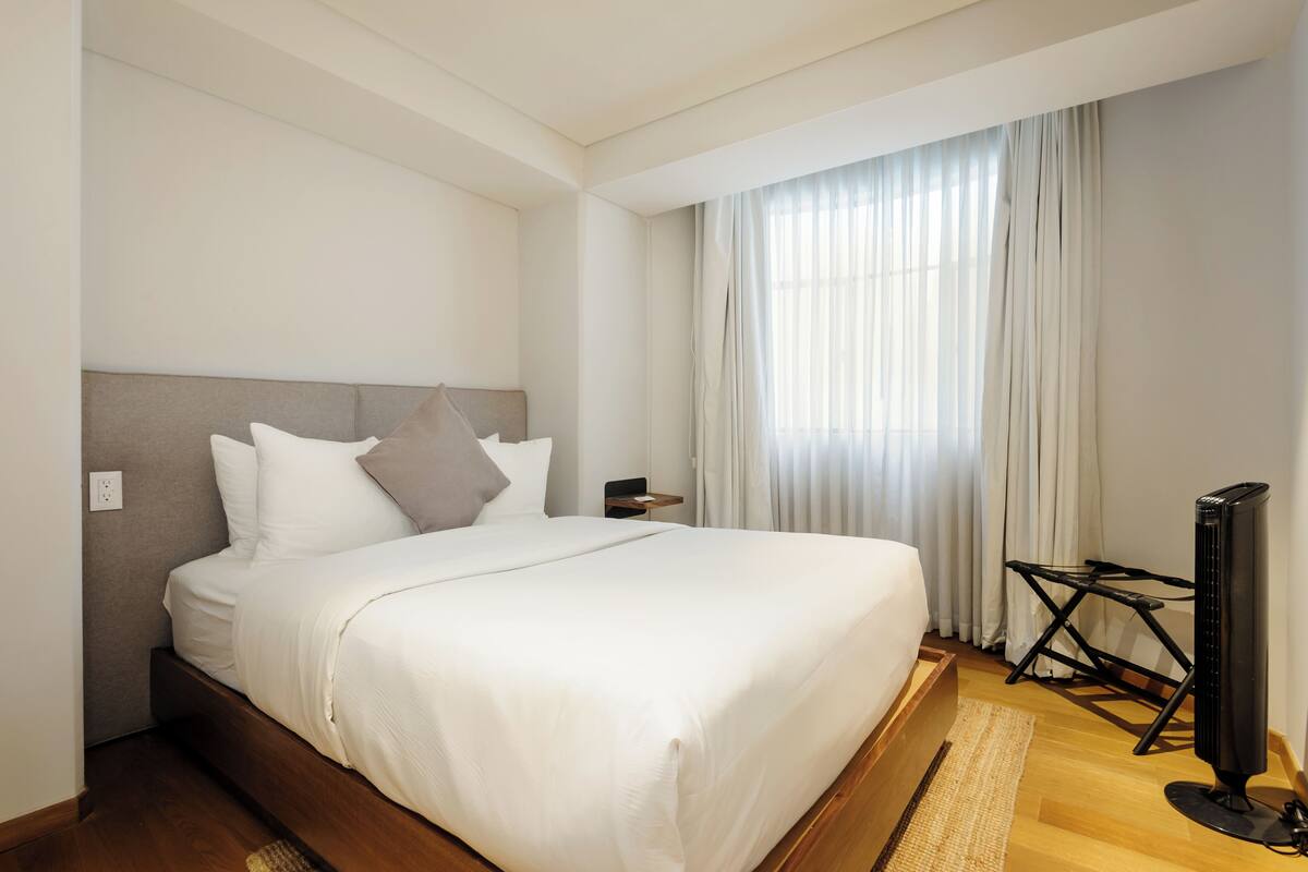 A neatly made bed with white linens, a bedside table, a flat-screen TV, and a window with sheer curtains.