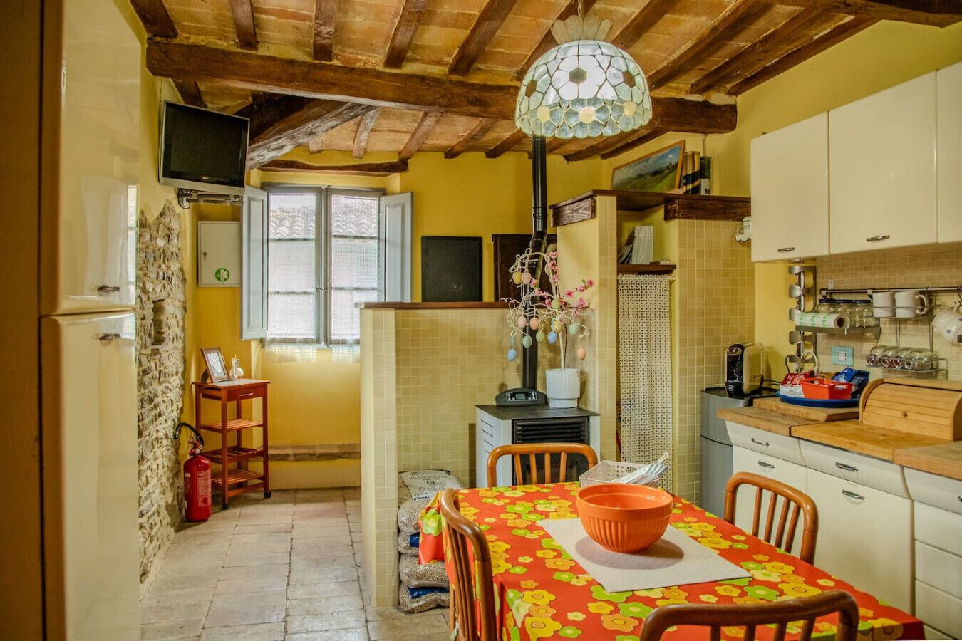 Une cuisine avec un plafond en bois, une table à manger recouverte d’une nappe à motifs floraux et une fenêtre avec des volets blancs.
