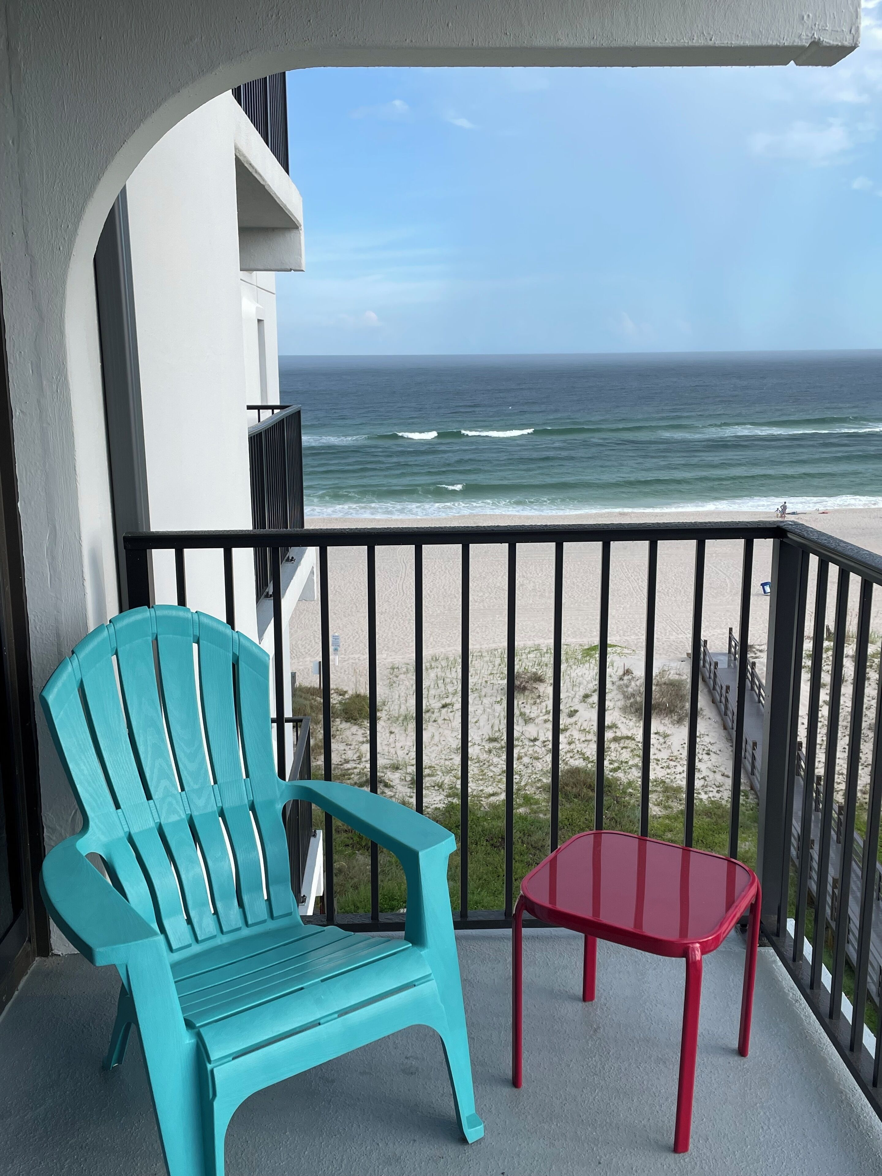 A balcony with a turquoise chair and a red table, overlooking a beach with waves and a clear sky.