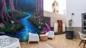 A courtyard with a mural of a forest stream, white chairs, a round table, and a potted plant. - Oakmoon House (Faro)