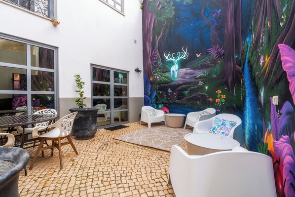 Un patio exterior con un mural de una escena boscosa, asientos al aire libre y piso de baldosas.