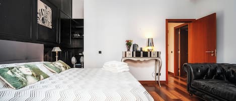 A bedroom with a bed, a black leather sofa, a desk with a lamp, and a door leading to another room.