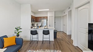 Un appartement moderne comprenant une cuisine, deux tabourets de bar, un canapé bleu avec un coussin jaune et un téléviseur situé à droite.