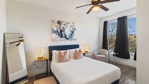 Une chambre d’hôtel comprenant un lit, deux tables de chevet avec des lampes, un ventilateur de plafond, un grand tableau abstrait, une chaise et une fenêtre avec des rideaux.