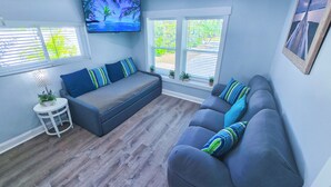 A living room with a grey sofa, a television mounted on the wall, and a window with blinds. - Walk to the Beach (Sarasota)