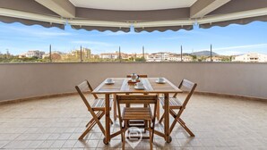 Een overdekt terras buiten met een tafel en stoelen, en gebouwen en heuvels op de achtergrond.
