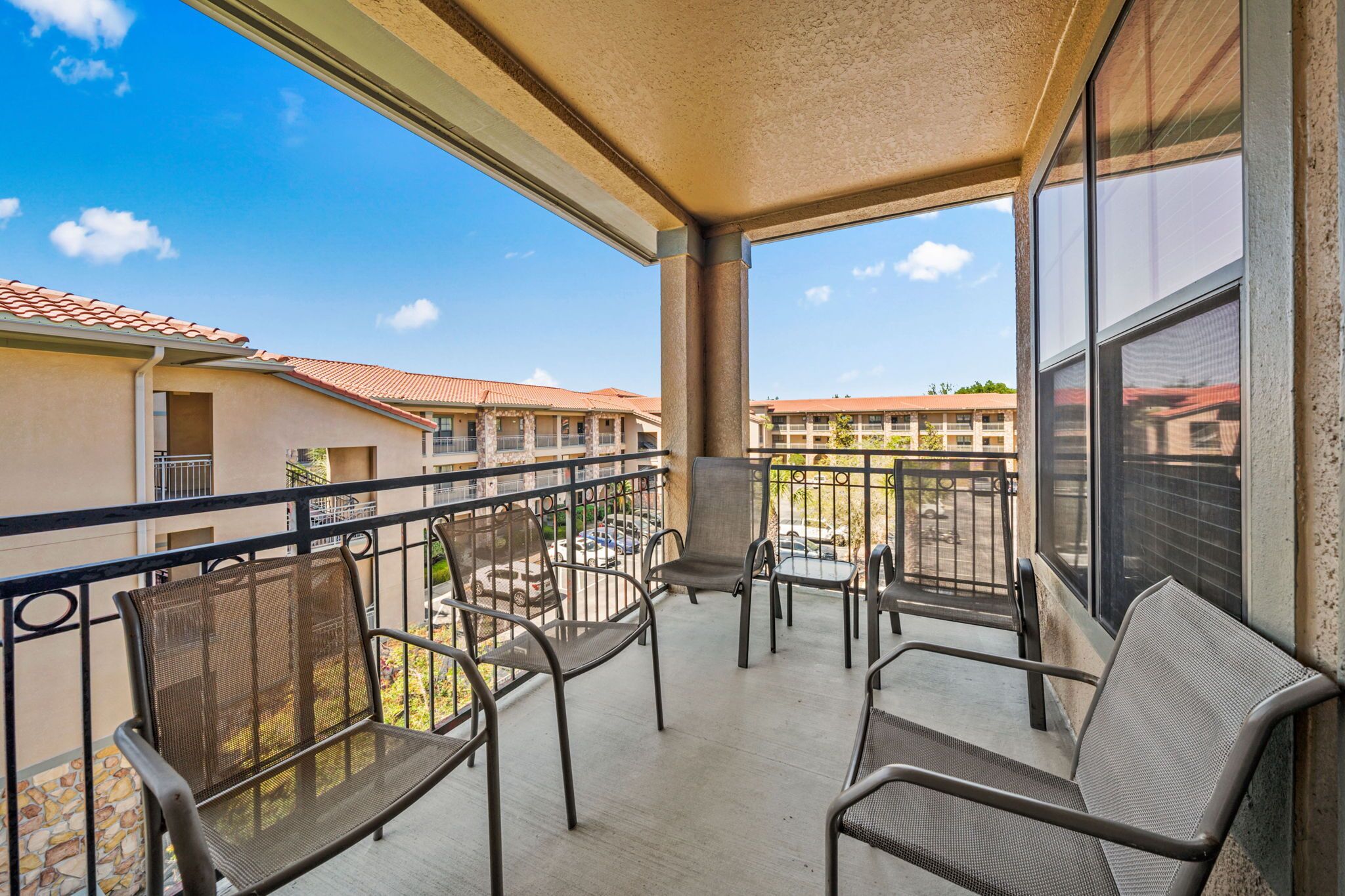 A balcony with metal chairs and a table, overlooking a courtyard with buildings in the background.