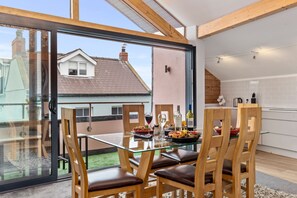 A dining area with a glass table and wooden chairs, a kitchen with white cabinets, and a view of a neighboring house through large glass doors. - Billycan Penthouse - Tenby Town Centre (Tenby)