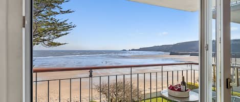 A balcony with a view of the beach, a table with a bottle, and a chair.