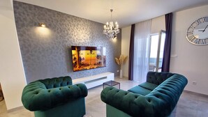 Un salon moderne avec deux canapés verts, un téléviseur à écran plat fixé au mur, un lustre et une grande horloge.