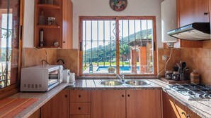 A kitchen with wooden cabinets, a microwave, a toaster, a sink, and a window with a view of a pool and greenery. - Finca Meraki Ojén by Ruralidays (Málaga)