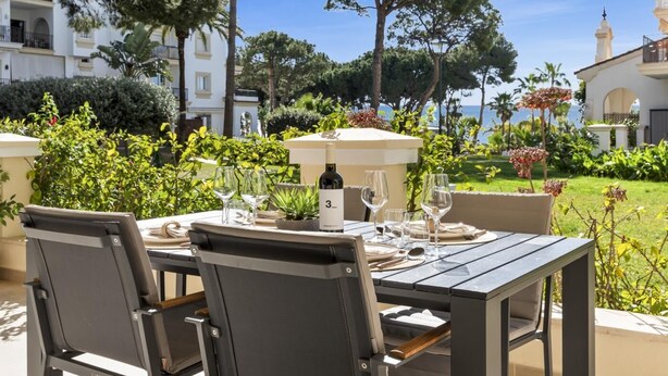 An outdoor dining area with a table set for two, surrounded by lush greenery and a view of the sea.