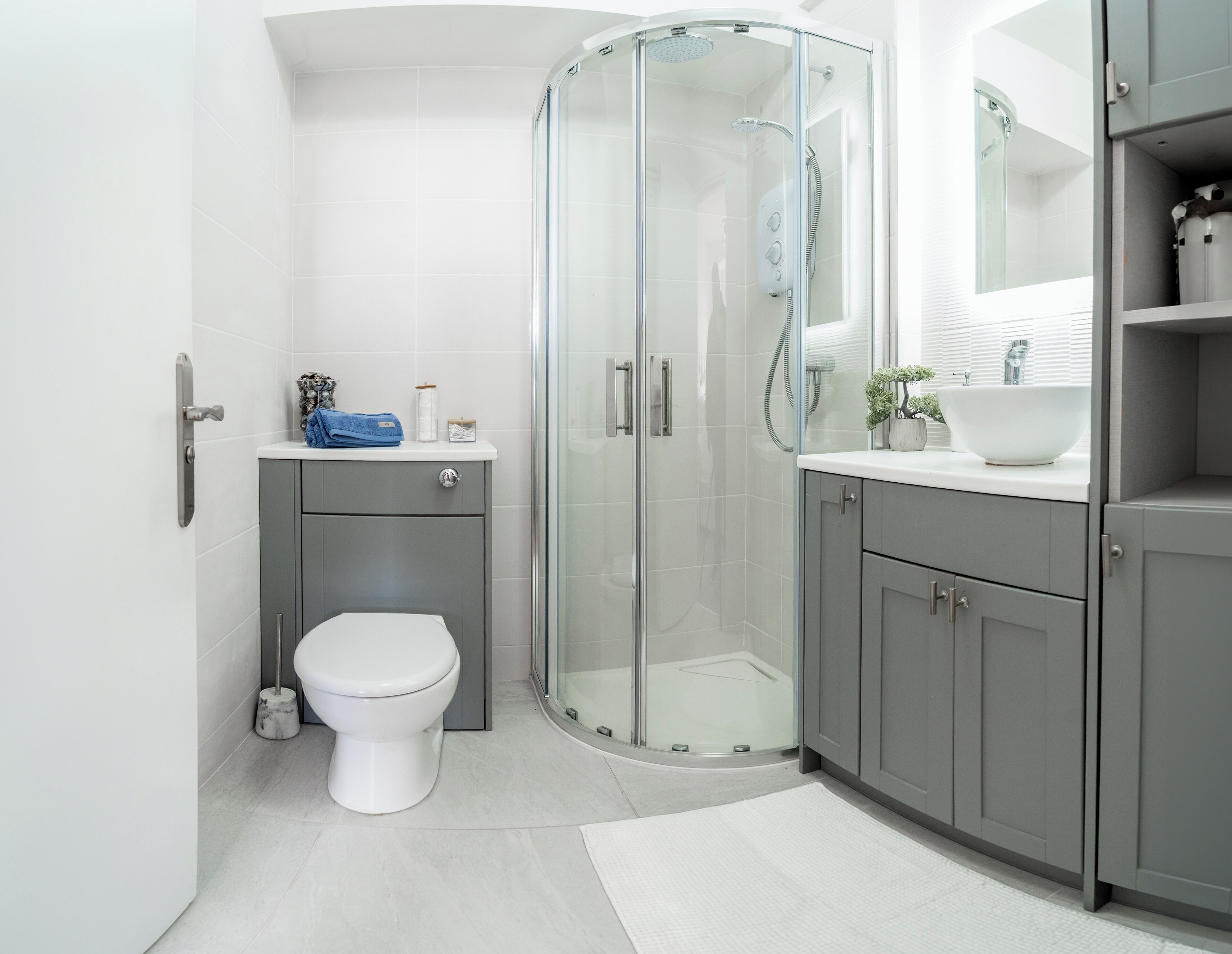 A modern bathroom with a glass-enclosed shower, a white toilet, grey cabinets, and a white sink with a mirror above.