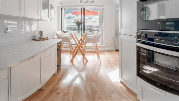 Une cuisine moderne avec des meubles blancs, une table à manger en bois et une vue sur la mer par une grande fenêtre.