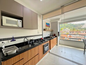 A modern kitchen with a microwave, stove, and oven. There is a balcony with a view of buildings and trees. - Apartment 109—new, cozy, 150 meters from the beach; we have other apartments available. (Ubatuba)