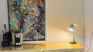 A room with a desk, a coffee machine, a chair, a ceiling fan, and a colorful painting on the wall.