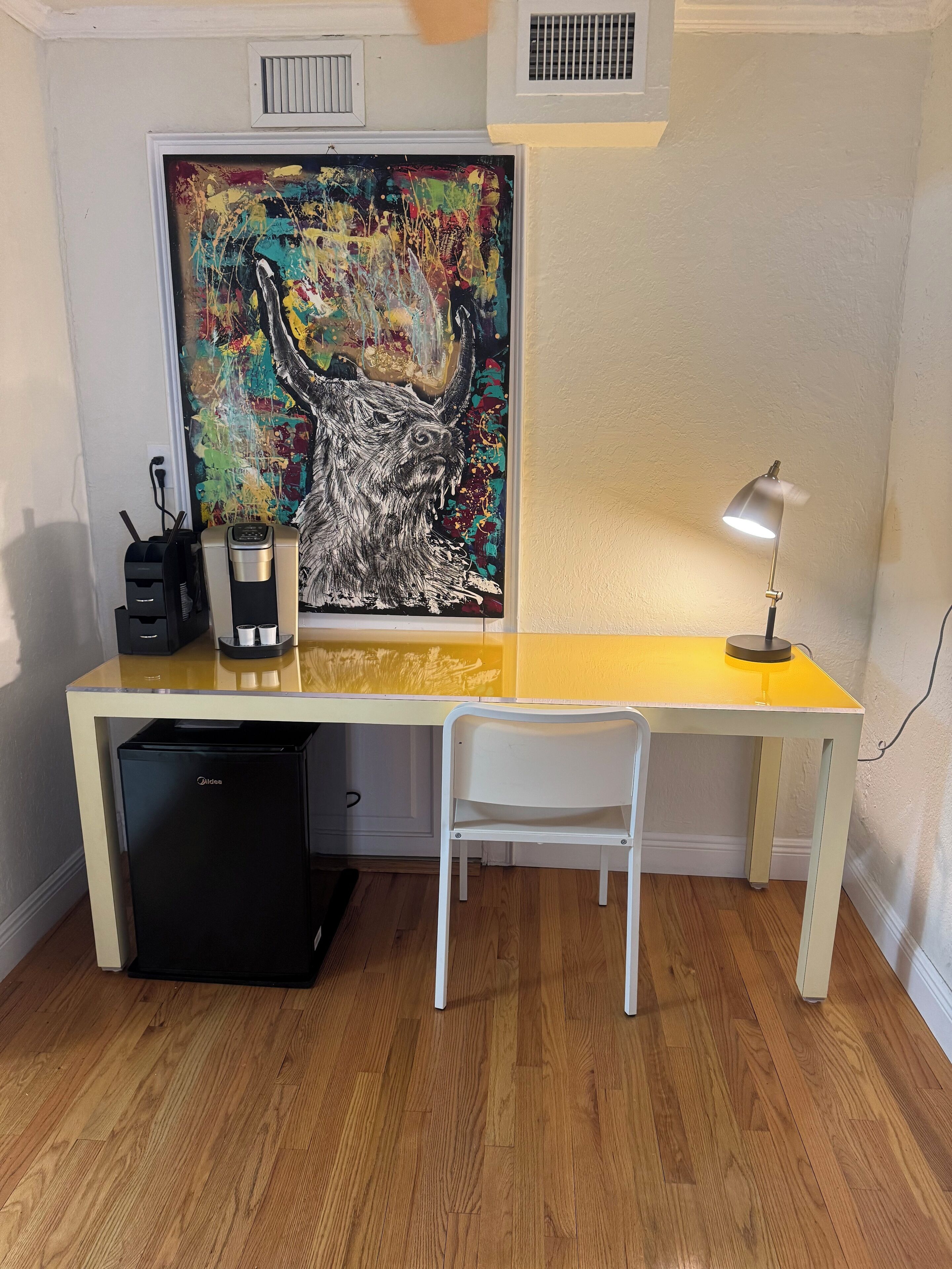 Un espacio de trabajo funcional con escritorio, silla, cafetera, refrigerador pequeño y un cuadro abstracto de grandes dimensiones.