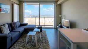 Una sala moderna con un sofá, una mesita y un televisor. Hay un balcón con vista a la playa y al océano.
