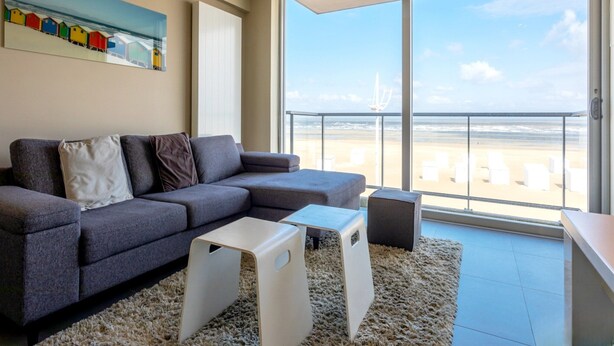 Una sala moderna con un sofá modular gris, una mesa de centro blanca y una mesita auxiliar pequeña también blanca. En la pared, un cuadro de casas coloridas, y la habitación tiene un gran ventanal corredizo que da a un balcón con vistas a la playa.