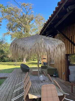 Una terraza de madera con mobiliario de mimbre, un parasol de paja y vistas a un jardín con césped y árboles.