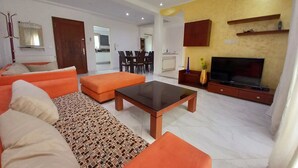 Una sala de estar moderna con un conjunto de sofás, una mesa de centro, un televisor y un comedor al fondo.