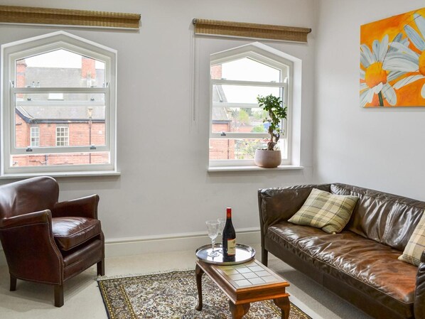 A living room with a brown leather sofa, a brown leather armchair, a small wooden coffee table with a tray holding a bottle and glasses, and a potted plant on a windowsill. - Ouse Retreat - sleeps 2 guests in 1 bedroom (York)