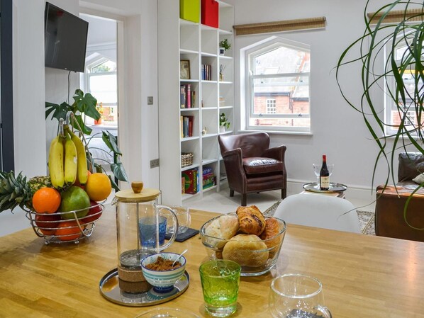 Un salon moderne avec une table en bois, un panier de fruits, des étagères, un fauteuil en cuir et une vue sur un bâtiment en briques, visible à travers une grande fenêtre.