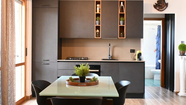Een moderne keuken met donkere kastjes, een glazen eettafel en een kleine potplant.