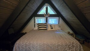 Ein Schlafzimmer mit schräg angesetzter Decke, einem Bett mit floralem Bettzeug, zwei Fenstern mit Vorhängen und einem kleinen Nachttisch.