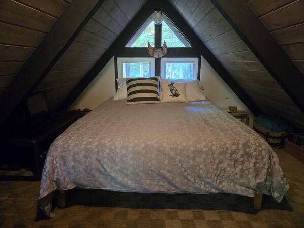 A bedroom with a sloped ceiling, a bed with floral bedding, two windows with curtains, and a small bedside table.
