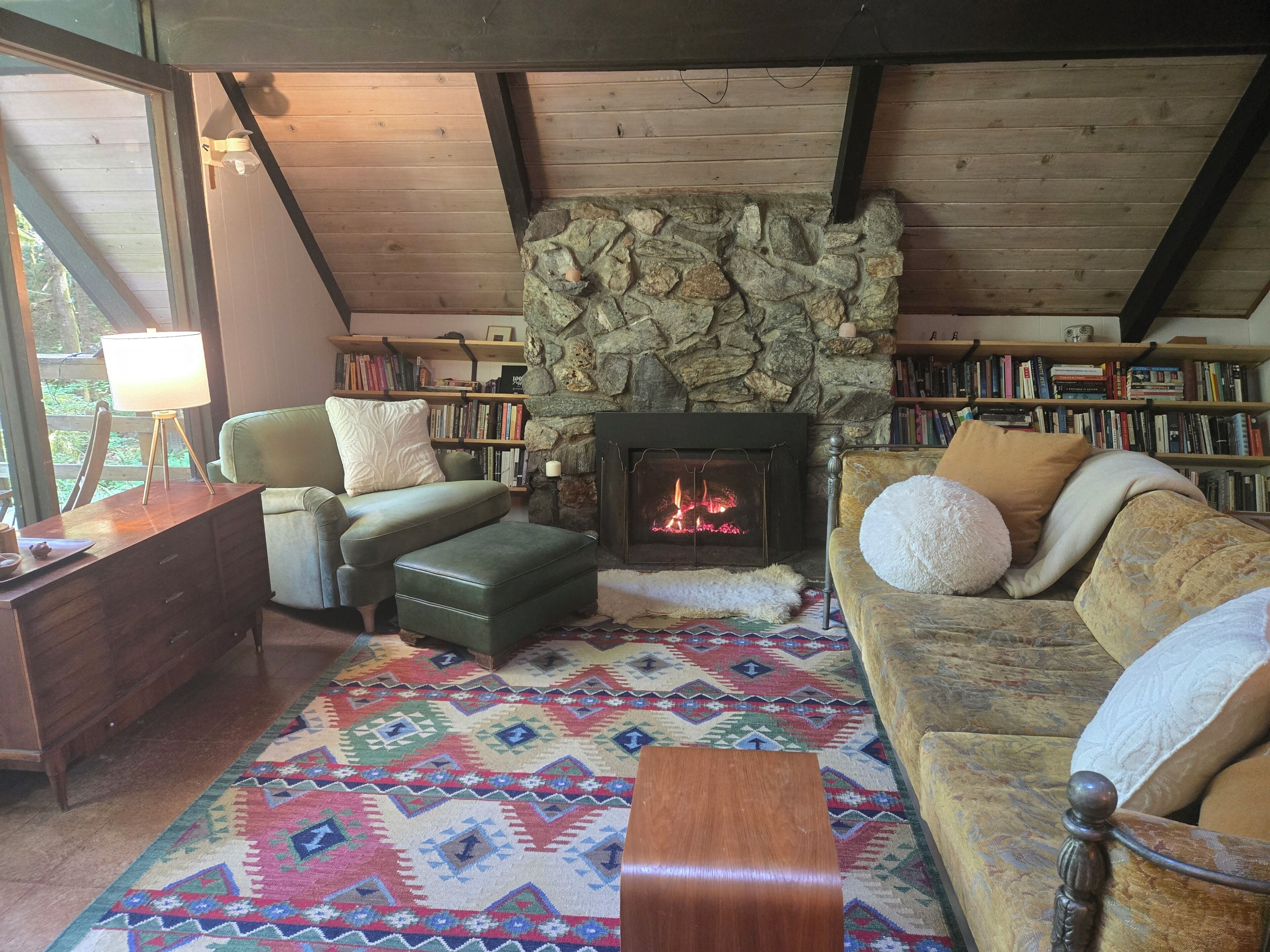 Un salon chaleureux avec une cheminée en pierre, un canapé vert, un tapis à motifs et une bibliothèque remplie de livres.