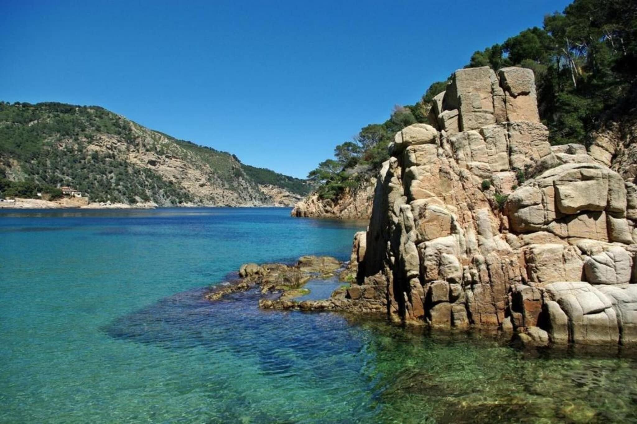 Eine Küstenlandschaft mit klarem, blauem Wasser, Felsformationen und einer hügeligen Kulisse.
