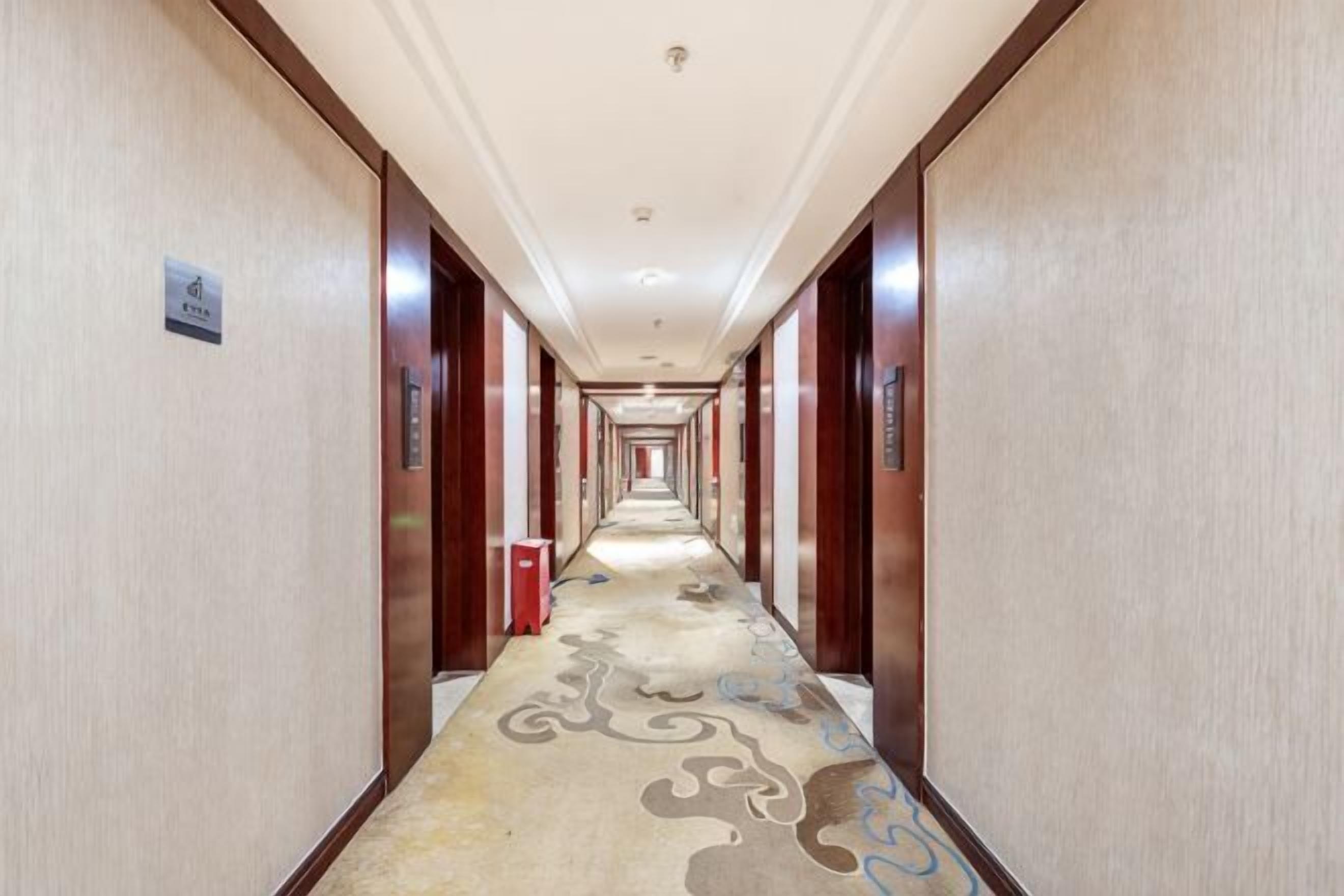 A long, narrow hotel corridor with a patterned carpet, wooden doors, and a red trash bin.