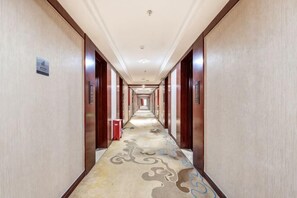 Um corredor de hotel comprido e estreito, com carpete estampado, portas de madeira e uma lixeira vermelha.