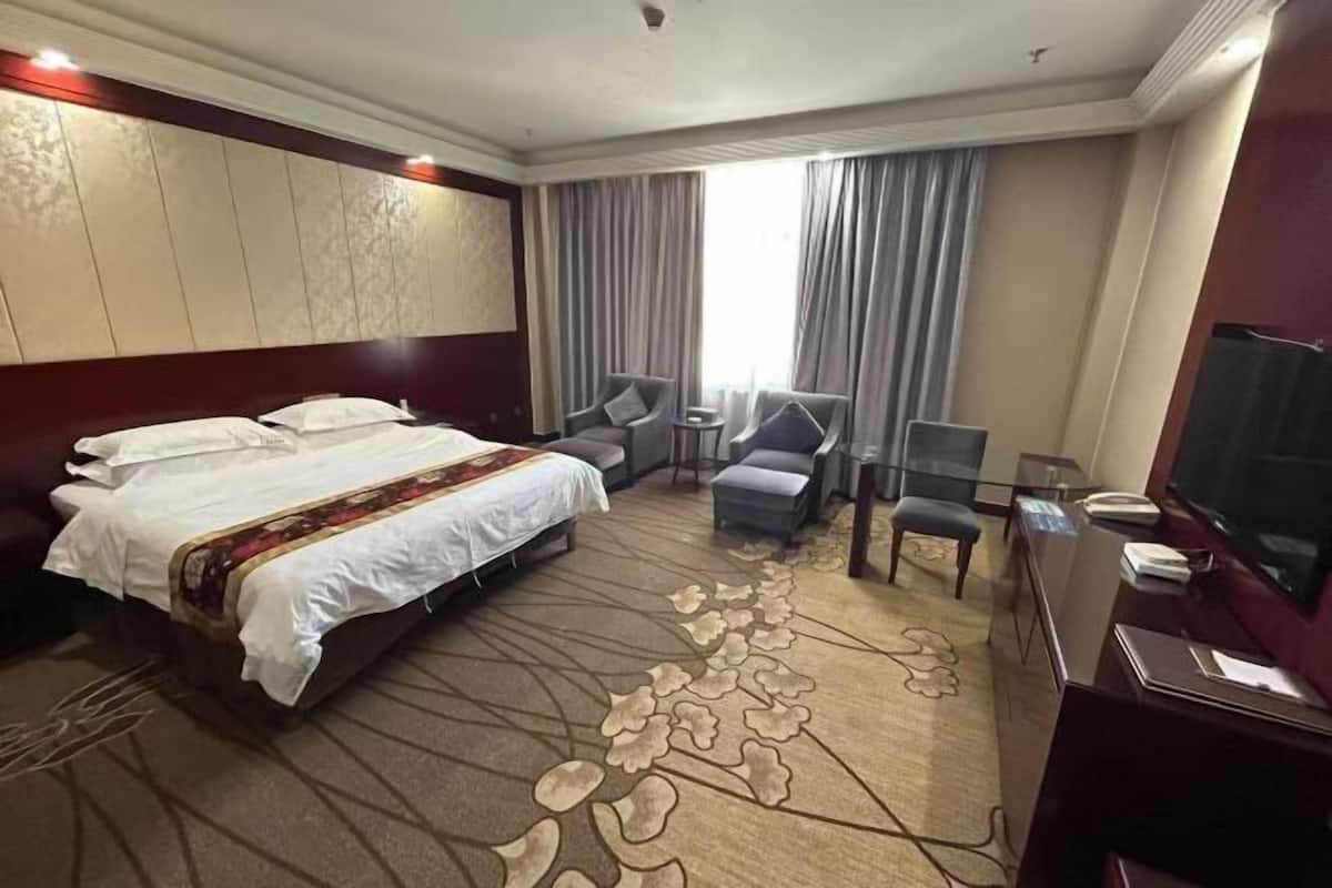 A hotel room with a large bed, a desk with a television, two armchairs, a small table, and a window with curtains.