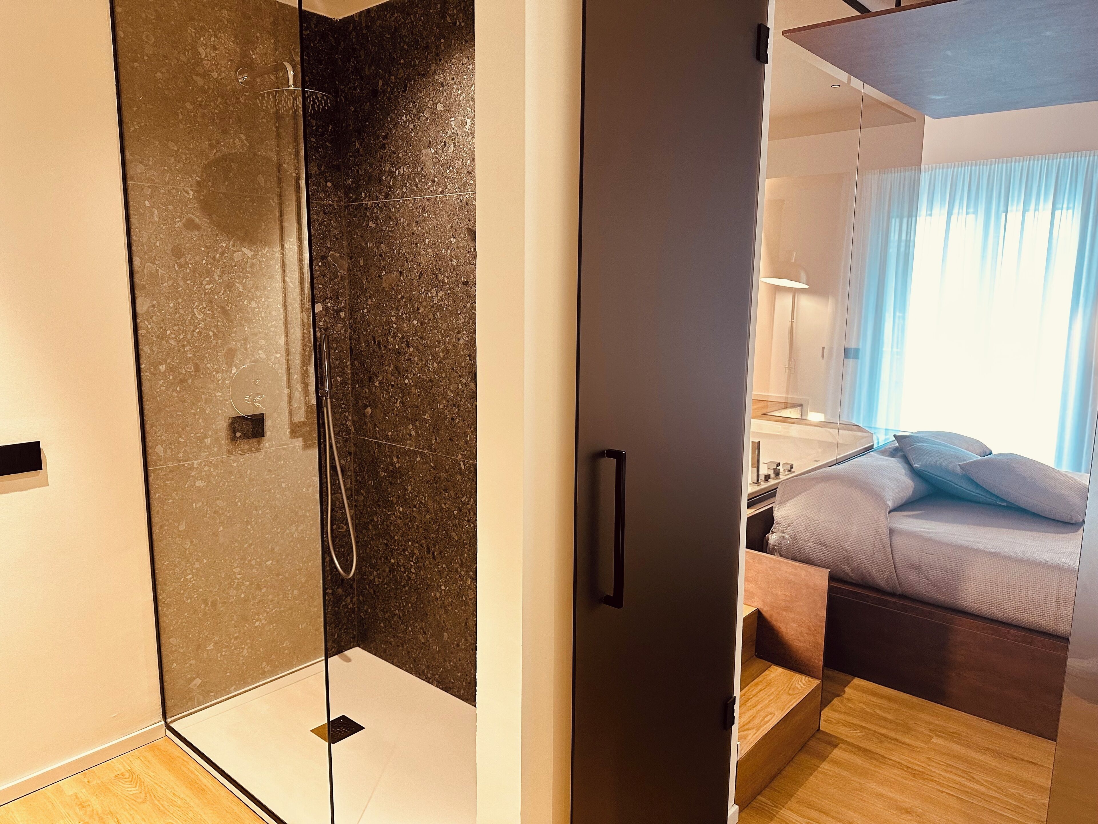 Une salle de bain moderne avec une douche en verre, un plancher en bois et un lit visible dans la pièce adjacente, agrémenté de coussins.