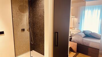 Une salle de bain moderne avec une douche en verre, un plancher en bois et un lit visible dans la pièce adjacente, agrémenté de coussins.