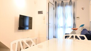 Una habitación con una mesa blanca, una silla blanca, un televisor de pantalla plana montado en la pared, una puerta con cortina y una cama con almohadas.