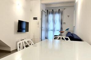A modern room with a flat-screen TV, a white table, a sofa, and a door leading to a balcony with curtains.