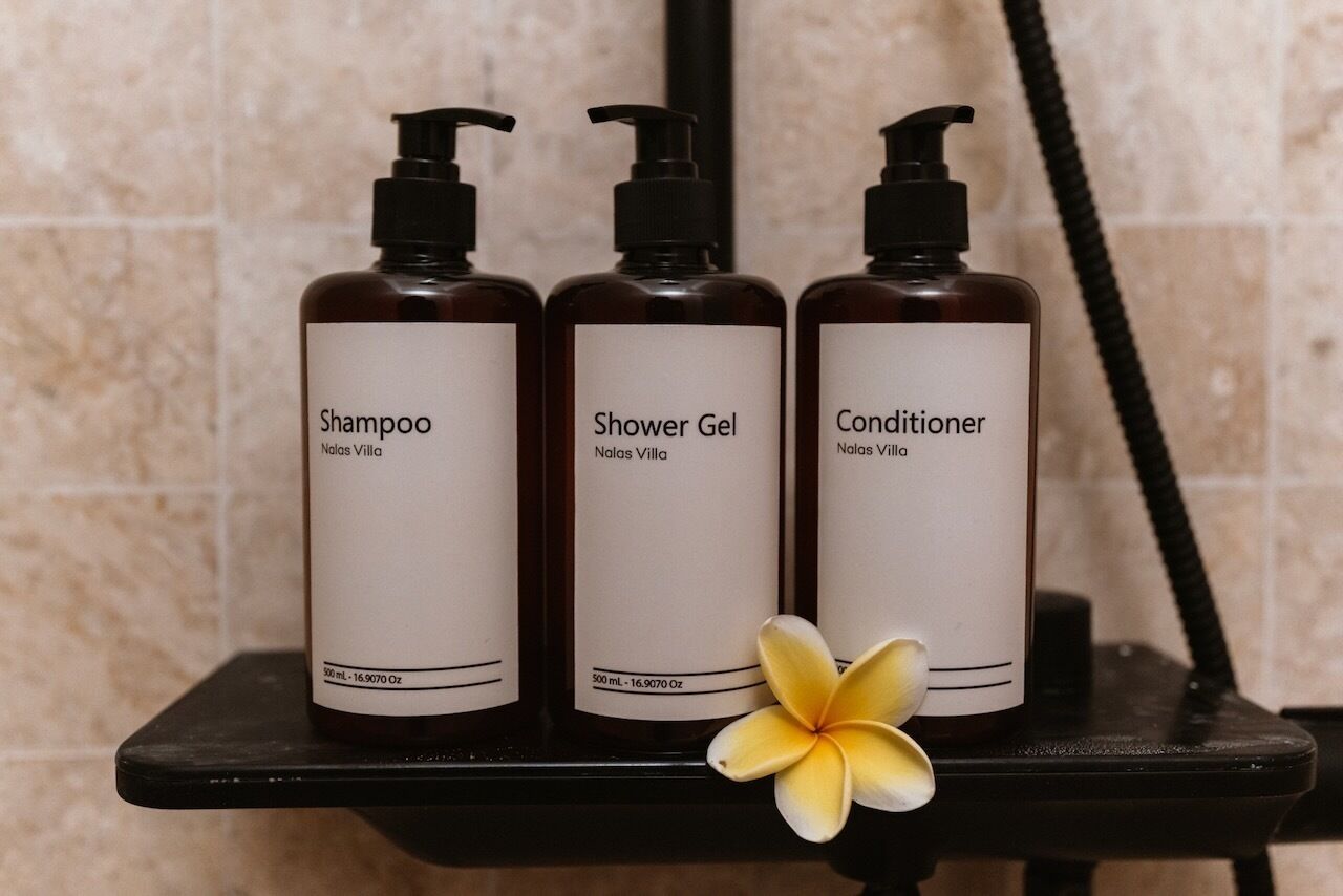 Three bottles of Nolas Villa products: Shampoo, Shower Gel, and Conditioner, on a black shelf with a single flower.