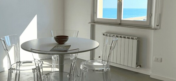 A round dining table with a bowl, clear chairs, a window with a view of the sea, and a white radiator. - M431 – Marcelli, new two-room apartment in the center facing the sea (Marcelli)