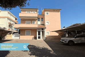 A multi-story building with a tiled roof, a car parked in the driveway, and a clear blue sky.