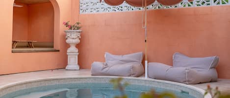 Piscina com guarda-sol rosa, duas espreguiçadeiras bege, um vaso decorativo branco e um banco de madeira.