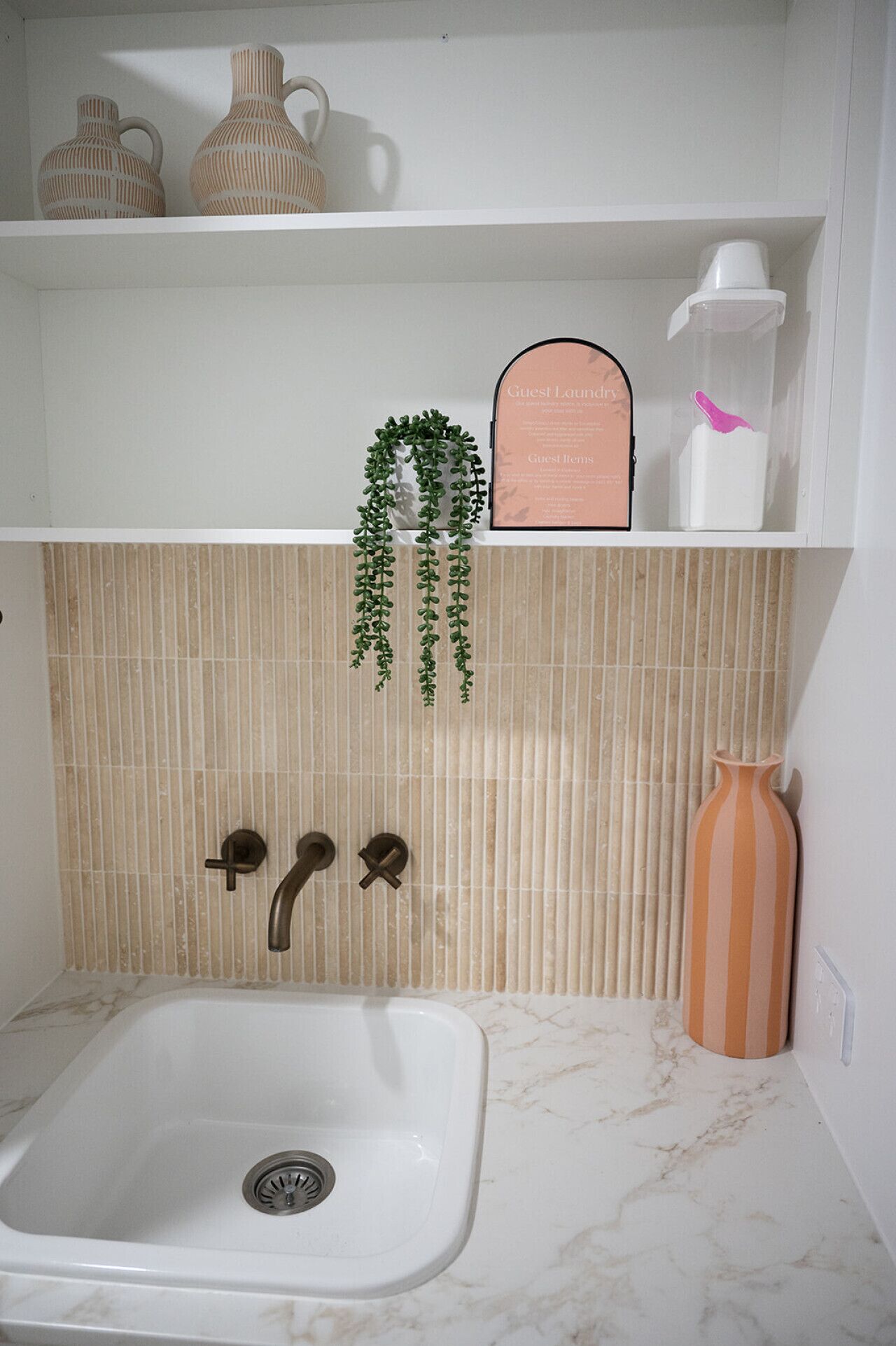 Un baño con un lavamanos blanco, dos grifos, un perchero con una planta, una estantería con adornos y un salpicadero de azulejos.