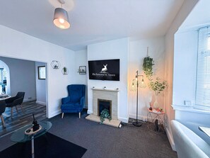 A modern living room with a fireplace, a blue armchair, a television mounted on the wall, and a dining area visible through an archway.