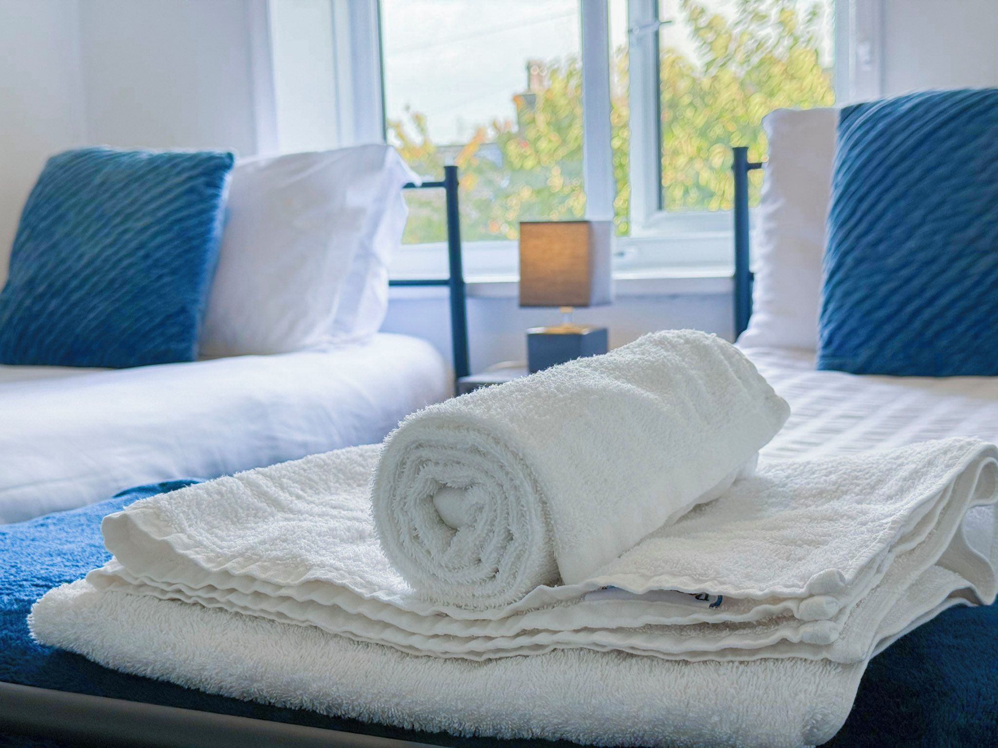 A neatly made bed with a rolled white towel and a folded white towel on a blue bedspread.