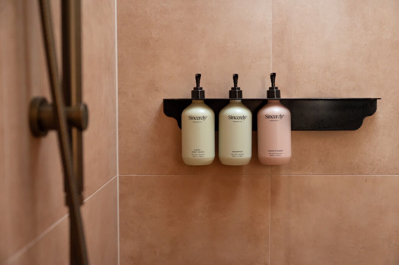 A bathroom with a black shelf holding three Sincerely Organic products: body wash, shampoo, and conditioner.