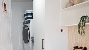 A compact laundry room with stacked washing machines, a sink, and a shelf with decorative items. - Pacific Sun Bargara ~ by the beach (Bargara)