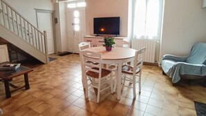 A dining area with a round table and chairs, a television on a stand, and a staircase leading upstairs. - Gîtes de France® - Gîte de Lacroux (LINAC)