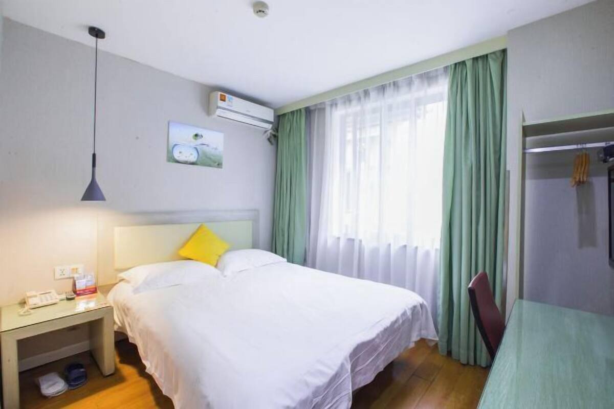 Una habitación de hotel con una cama, una mesita de noche con teléfono y lámpara, una ventana con cortinas y un equipo de aire acondicionado.